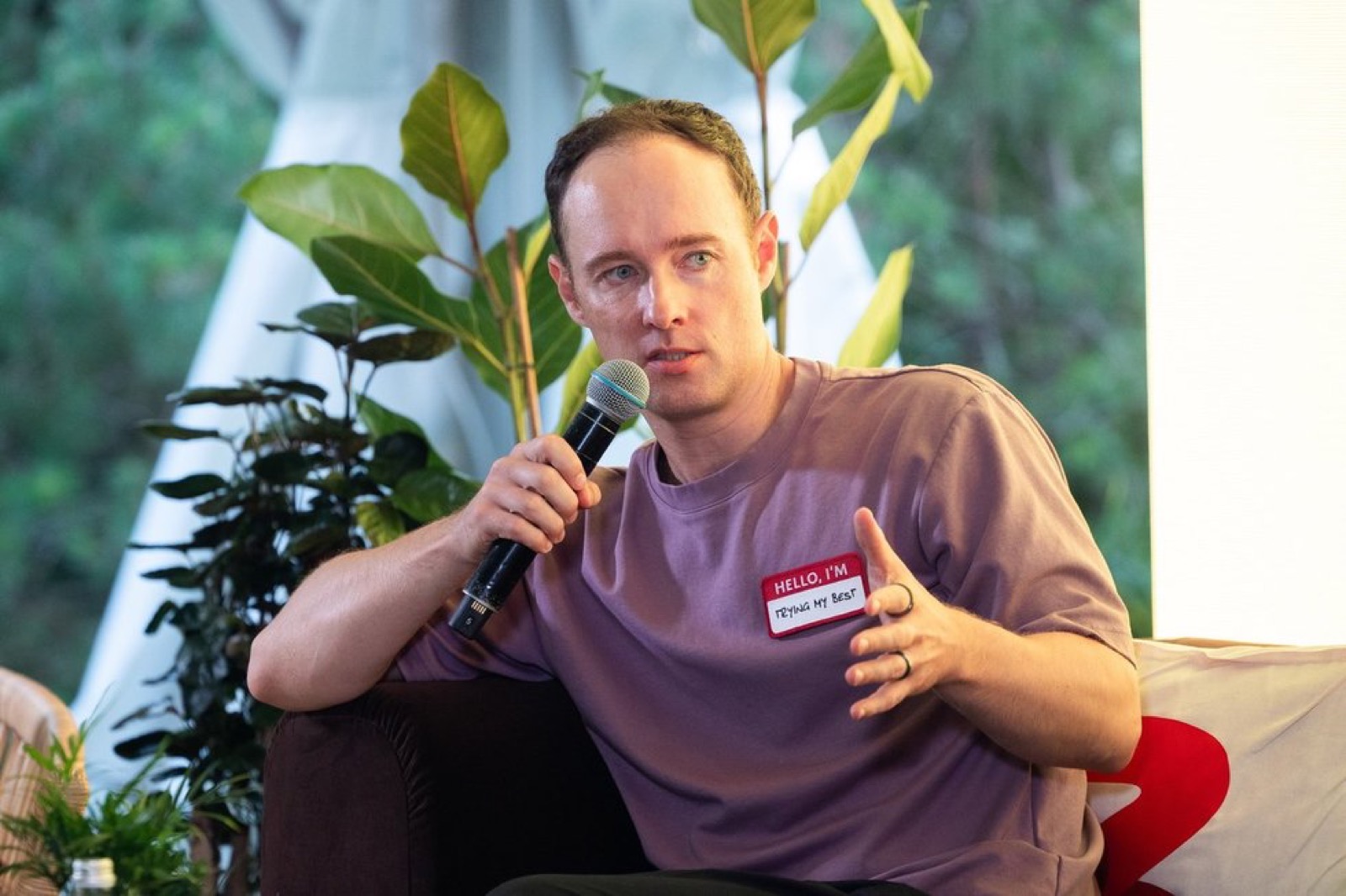 Martynas Kairys holding a microphone on a couch at the Būtent! festival 2025, name tag reading 'trying my best'