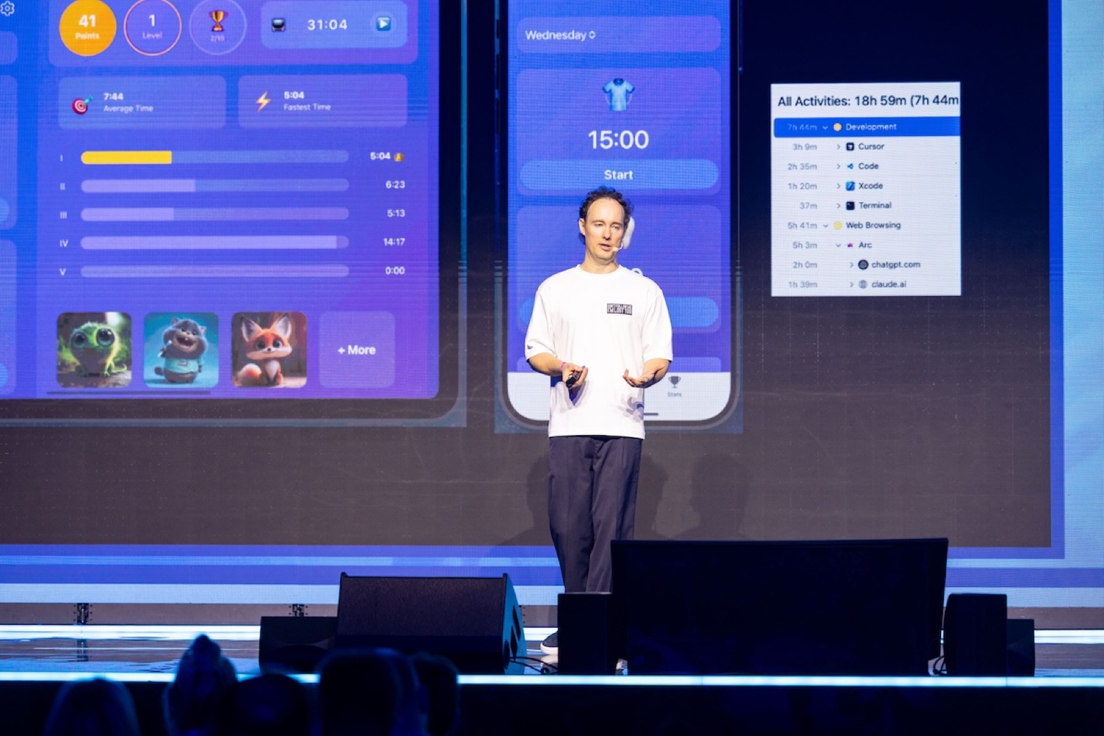 Martynas Kairys on the LOGIN 2025 stage, white t-shirt, presenting app screenshots on the big screens behind him
